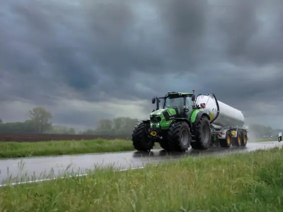En grønn Deutz-Fahr Series 7 traktor kjører på en våt vei med en stor tankvogn.