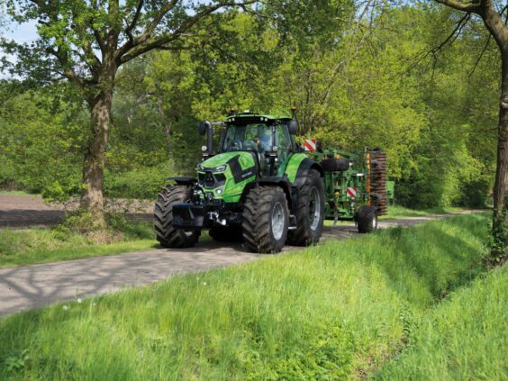 Grønn Deutz-Fahr Series 7 traktor med landbruksutstyr på en vei omgitt av trær.