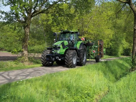 Grønn Deutz-Fahr Series 7 traktor med landbruksutstyr på en vei omgitt av trær.