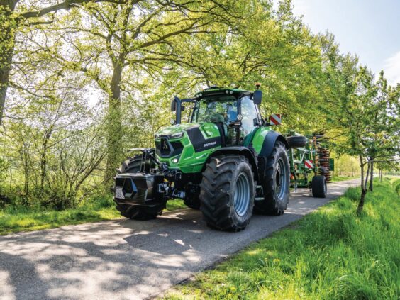 Grønn Deutz-Fahr Series 7 traktor som kjører på en vei mellom frodige, grønne trær.