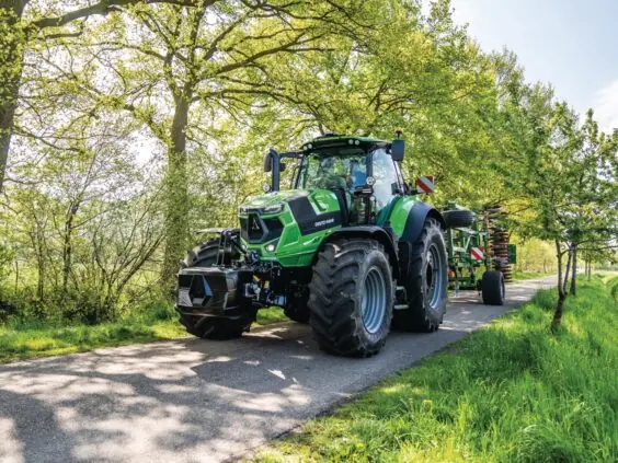 Grønn Deutz-Fahr Series 7 traktor som kjører på en vei mellom frodige, grønne trær.