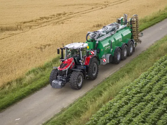 Rød traktor trekker en SAMSON PG II Genesis gjødselvogn på en vei ved åkeren.