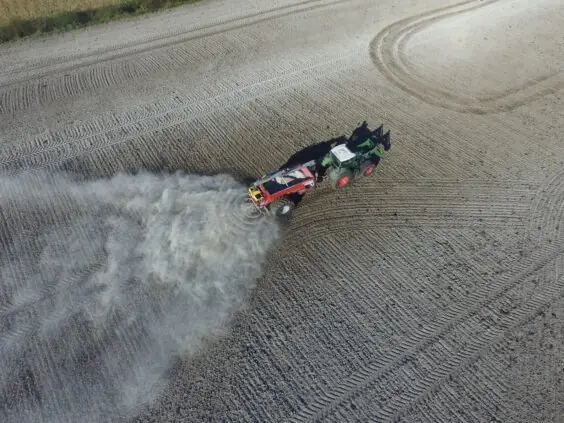 En grønn traktor trekker en rød Bredal kalkspreder som sprer kalk på en åker.