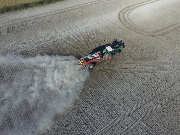 En grønn traktor trekker en rød Bredal kalkspreder som sprer kalk på en åker.