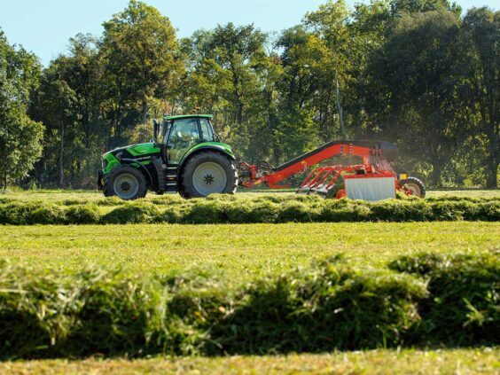 Grønn Deutz-Fahr Series 6 traktor i arbeid på en gresskledd åker med skog i bakgrunnen.