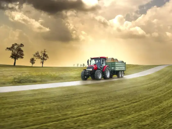 En rød traktor som trekker en grønn tilhenger med høyballer på en landevei.