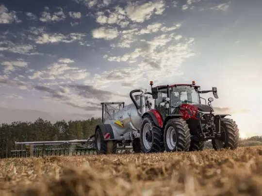 Rød traktor med gjødselvogn på en åker under en skyete himmel ved solnedgang.