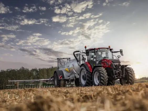 Rød traktor med gjødselvogn på en åker under en skyete himmel ved solnedgang.