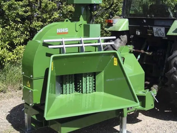 En grønn NHS 220cv flishugger montert bak på en traktor utendørs.