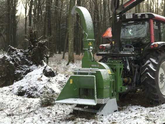 En grønn NHS 220ec flishugger montert på en traktor i et snødekket skogslandskap.