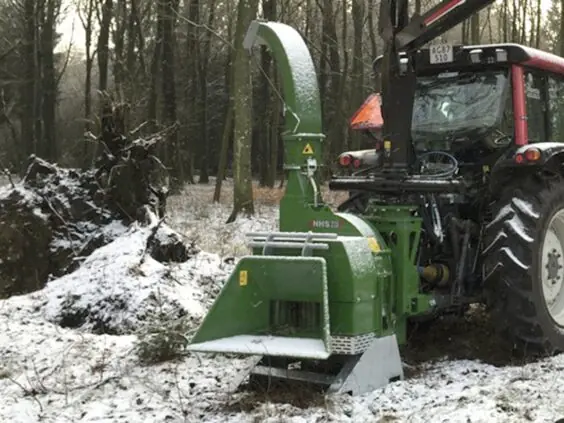 En grønn NHS 220ec flishugger montert på en traktor i et snødekket skogslandskap.