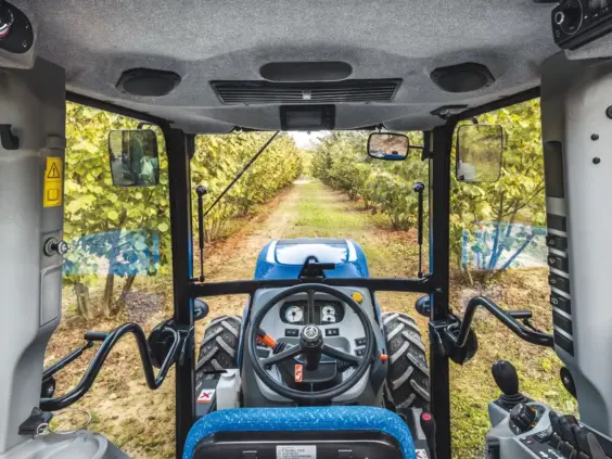 Innvendig utsikt fra førerhuset i en New Holland T4 som kjører mellom rader med frukttrær.