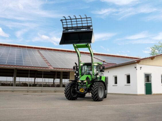 Grønn Deutz-Fahr Series 5 traktor med frontlaster hevet foran en driftsbygning med solcellepaneler.