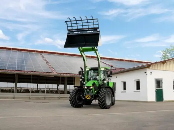 Grønn Deutz-Fahr Series 5 traktor med frontlaster hevet foran en driftsbygning med solcellepaneler.