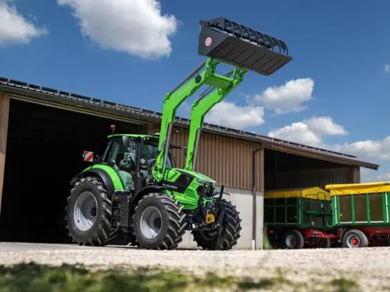 En grønn Deutz-Fahr Series 7 traktor med frontlaster løftet høyt foran en låve.