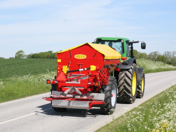 En grønn traktor som trekker en rød Bredal kalkspreder på en asfaltert vei.
