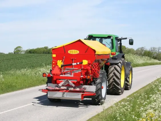 En grønn traktor som trekker en rød Bredal kalkspreder på en asfaltert vei.