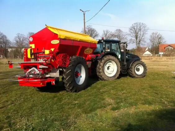 Rød Bredal kalkspreder med gult deksel koblet til en traktor på et grønt jorde.