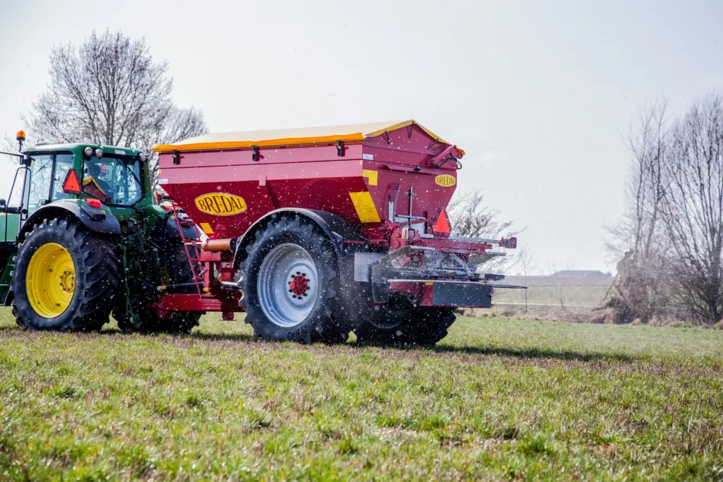 Grønn traktor som trekker en rød Bredal kalkspreder i arbeid på en grønn åker.