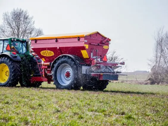 Grønn traktor som trekker en rød Bredal kalkspreder i arbeid på en grønn åker.