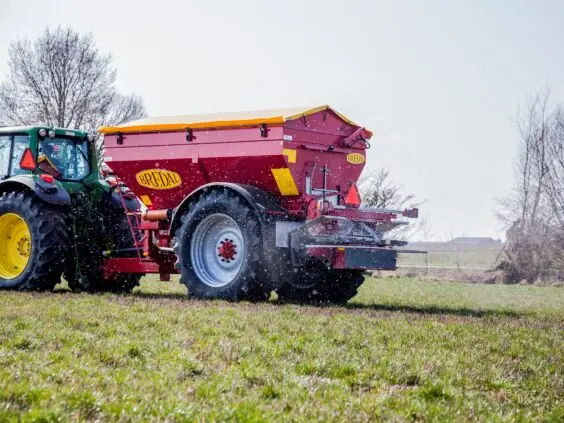 En rød Bredal kalkspreder trekkes av en grønn traktor på et grønt jorde.