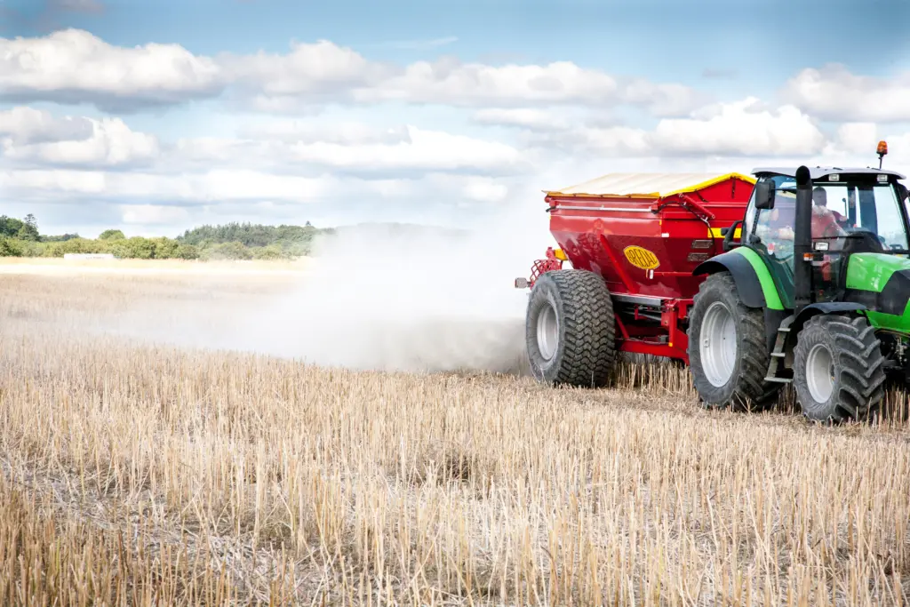 En grønn traktor trekker en rød Bredal kalkspreder som sprer kalk på en åker.