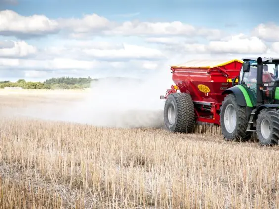 En grønn traktor trekker en rød Bredal kalkspreder som sprer kalk på en åker.