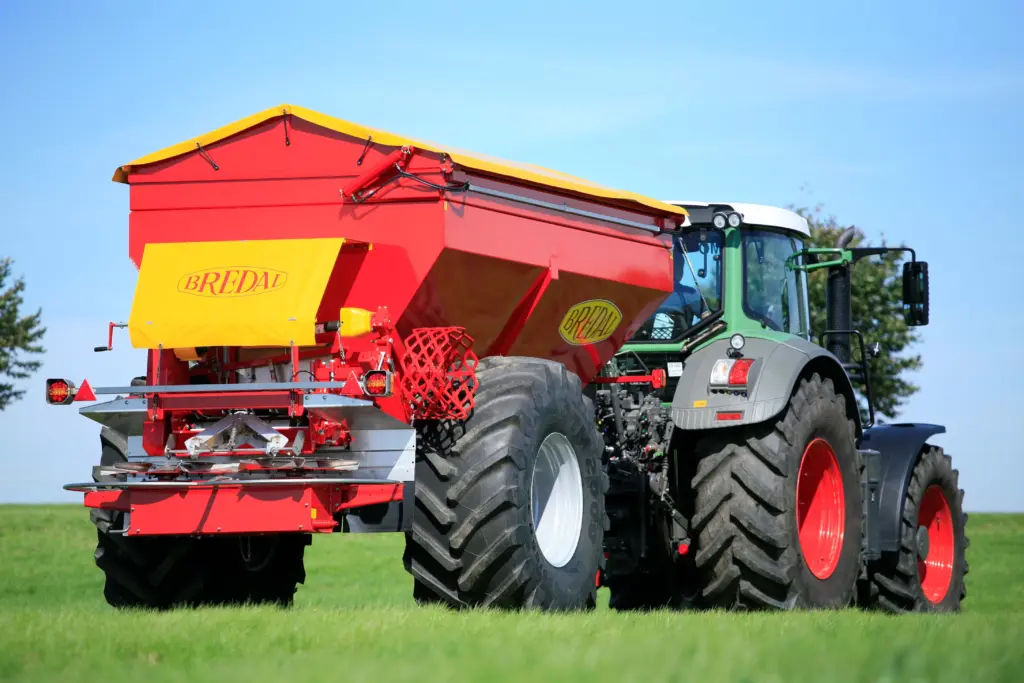 En rød Bredal kalkspreder montert bak en grønn traktor på et grønt jorde.