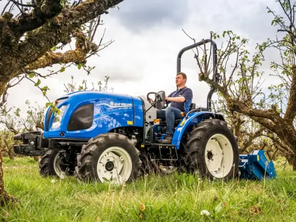 En blå New Holland mini traktor Boomer 25 C-55 kjøres gjennom en frukthage.