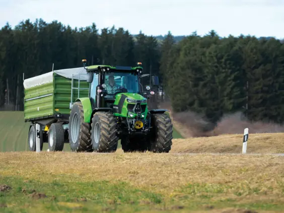 Grønn Deutz-Fahr Series 6C traktor som trekker en tilhenger langs en landevei ved en skog.