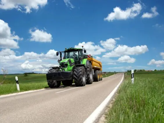 Grønn Deutz-Fahr Series 7 traktor med gul tilhenger kjører på en solfylt landevei.