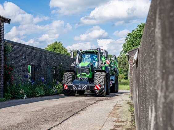 En grønn Deutz-Fahr Series 9 traktor som kjører mellom gårdsbygninger under en blå himmel.