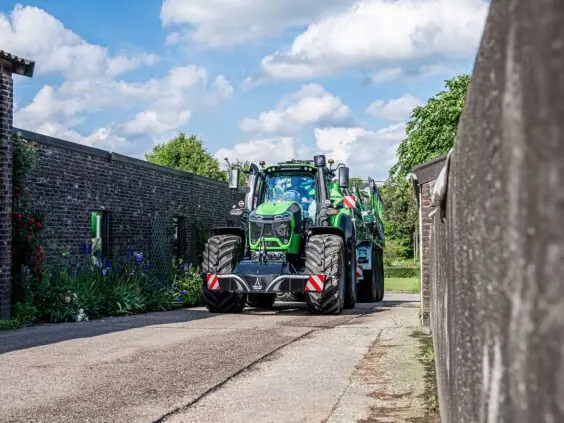 En grønn Deutz-Fahr Series 9 traktor som kjører mellom gårdsbygninger under en blå himmel.