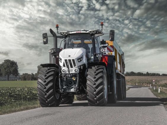Hvit Steyr Cervus CVT traktor med tilhenger på vei gjennom et åpent landskap under skyer.