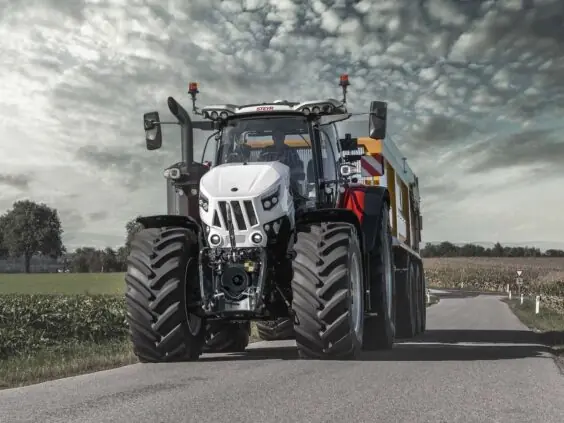 Hvit Steyr Cervus CVT traktor med tilhenger på vei gjennom et åpent landskap under skyer.