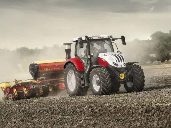 Rød og hvit Steyr Profi traktor i arbeid med såmaskin på en støvete åker.