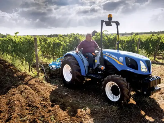 Blå New Holland traktor fra T3F-serien i arbeid i en vingård under skyet himmel.