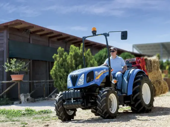 Blå New Holland traktor fra T3F-serien kjører med høyballer på en gård.