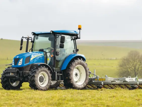 En blå New Holland T4S traktor arbeider på en grønn eng med landbruksutstyr bak.