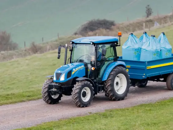 Blå New Holland T4S traktor med henger lastet med gjødselsekker på en grusvei.