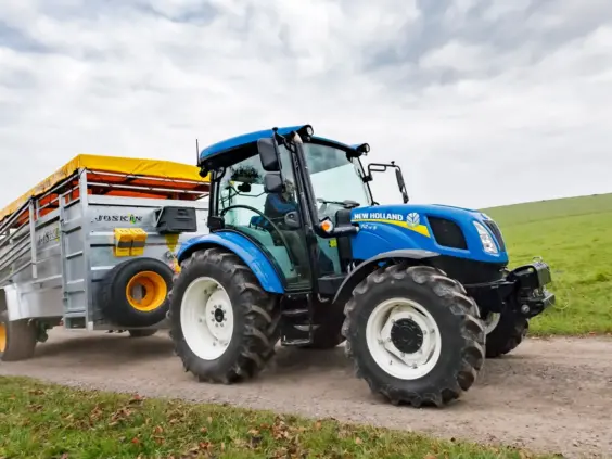En blå New Holland T4S traktor som trekker en Joskin tilhenger på en landevei.