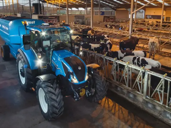 Blå New Holland T5 Electro Command traktor som fôrer kuer i et stort, moderne fjøs.