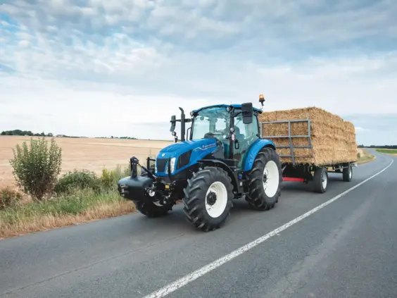 Blå New Holland T5 Utility traktor som transporterer høyballer på en asfaltert landevei.