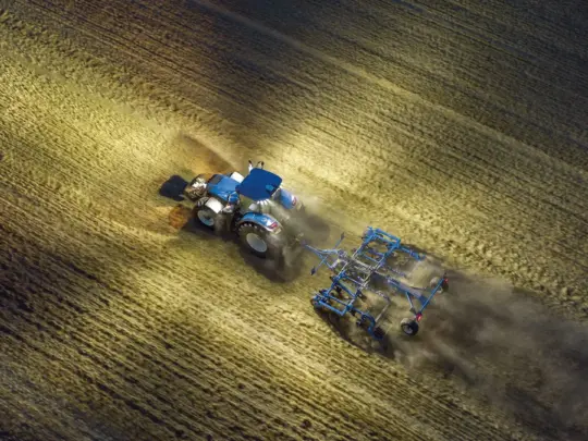 En blå T7 Heavy Duty traktor som arbeider på en åker i kveldsmørket.