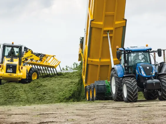 Blå New Holland T7 SWB traktor ved siden av en gul tilhenger som tipper gress.