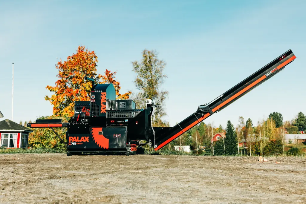 Palax C751 Smart vedmaskin med hevet utmatingsbånd, plassert på en grusplass med høstfarget løvtre og rødt våningshus i bakgrunnen.