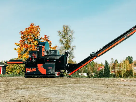 Palax C751 Smart vedmaskin med hevet utmatingsbånd, plassert på en grusplass med høstfarget løvtre og rødt våningshus i bakgrunnen.