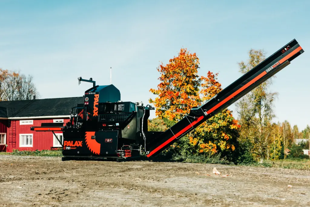 Palax C751 Smart vedmaskin sett fra siden foran en rød låve, med utmatingsbåndet hevet i arbeidsstilling.