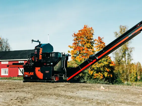Palax C751 Smart vedmaskin sett fra siden foran en rød låve, med utmatingsbåndet hevet i arbeidsstilling.