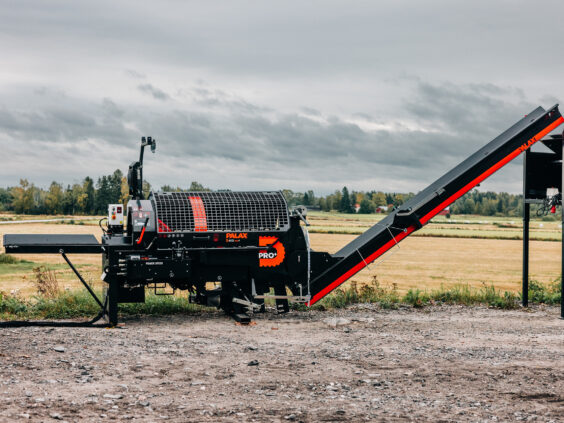 En Palax D410 vedmaskin står utendørs på en grusplass med et jorde i bakgrunnen.